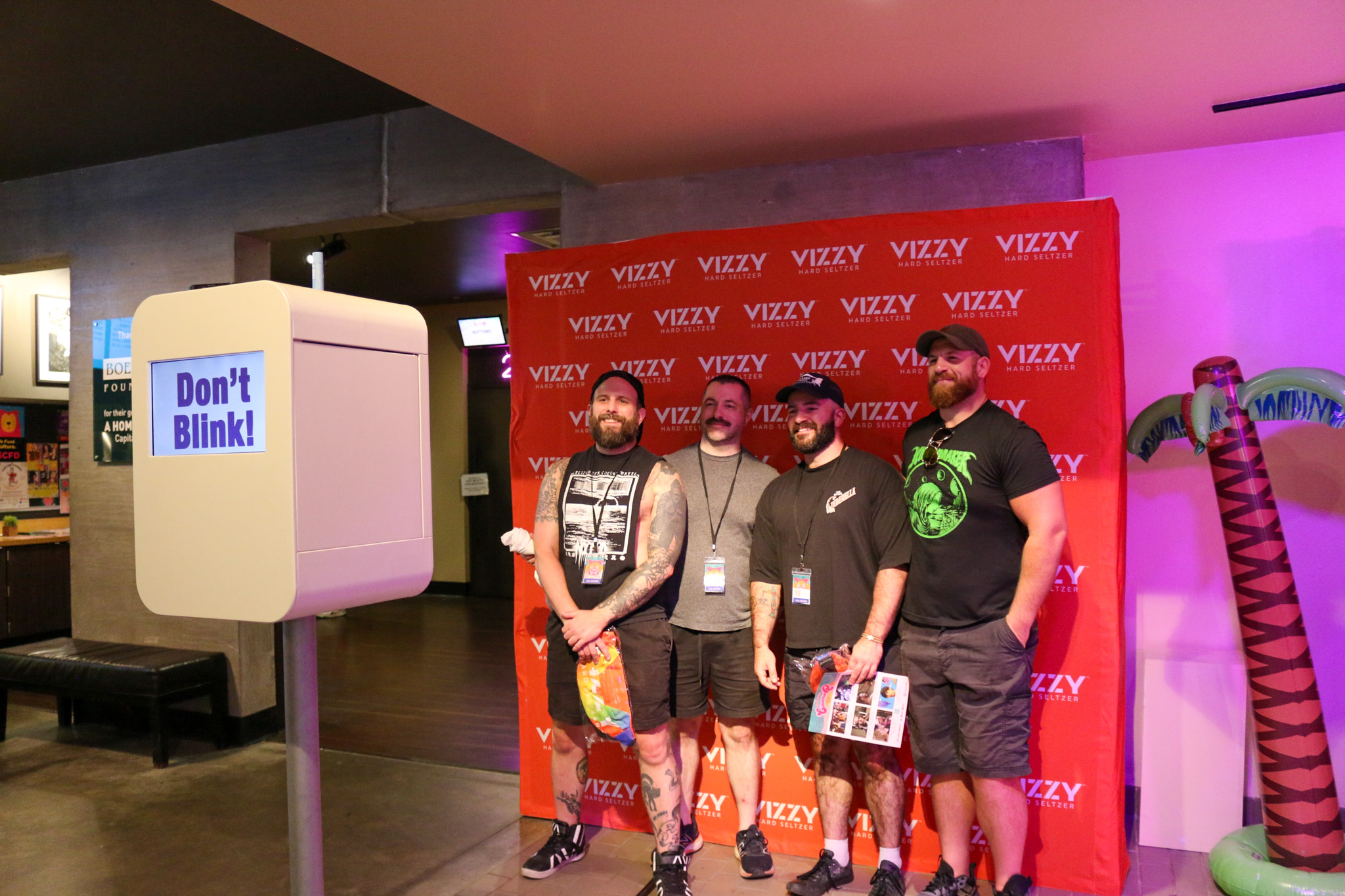 Four men standing before a red Vizzy backdrop, smiling near a "Don't Blink!" photo booth