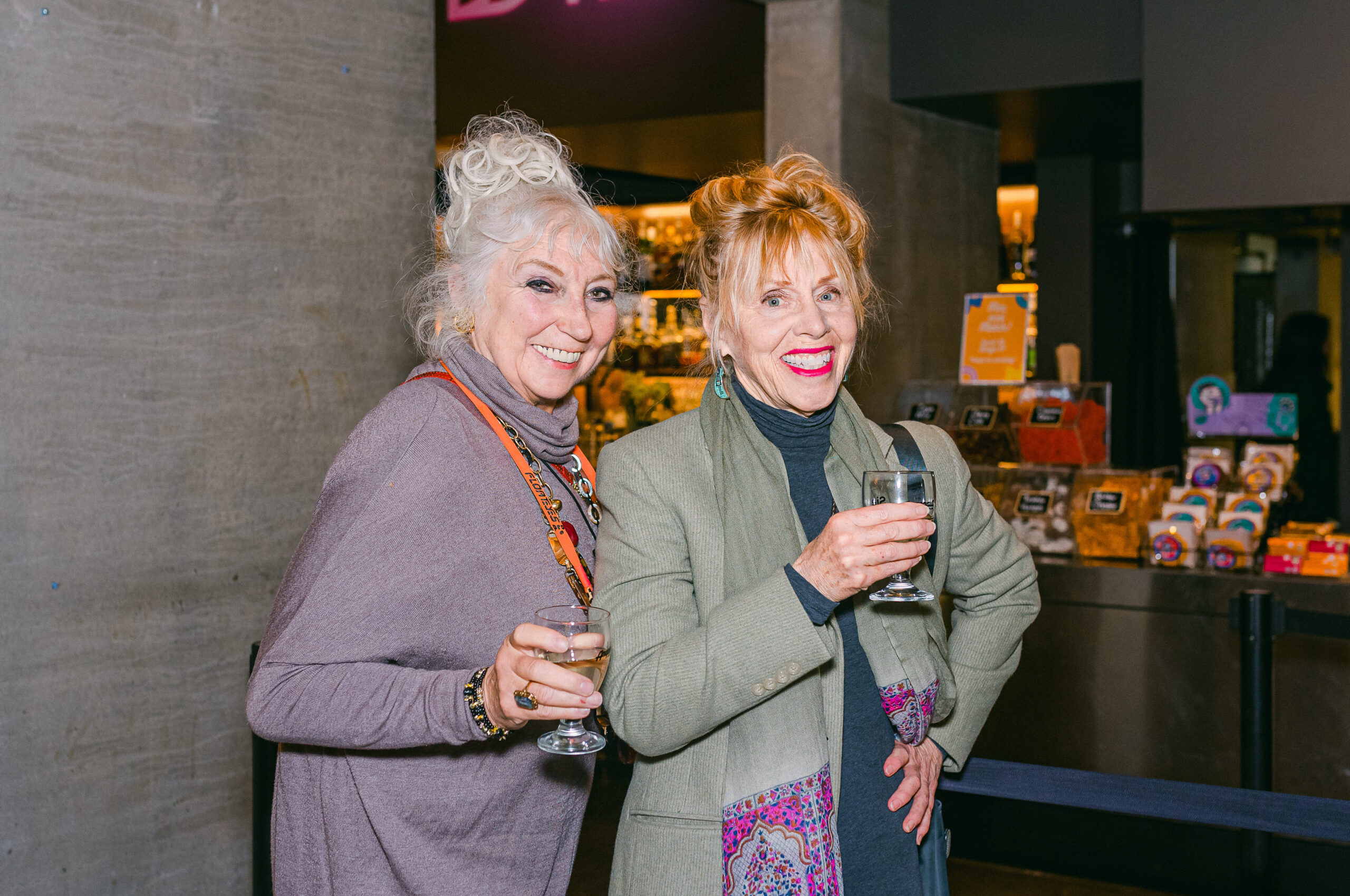 [gpt-4o]: Two women smiling, holding drinks at an indoor event, standing beside a counter with snacks