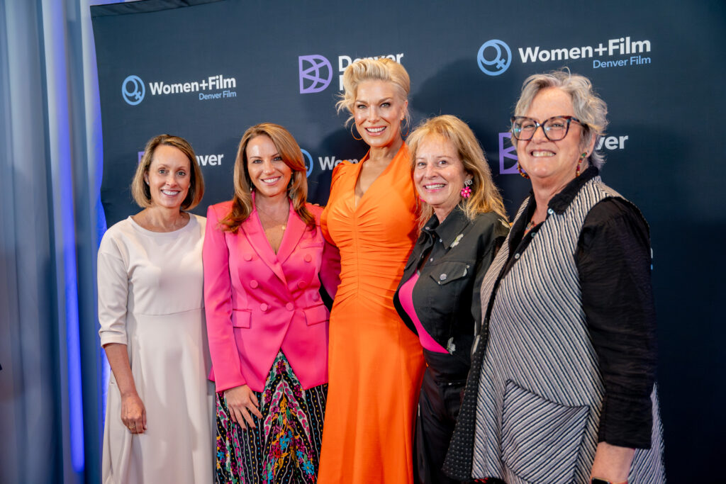 Five women smiling at Women+Film Denver Film event backdrop, including Premiere-Circle-24 in a bright orange dress