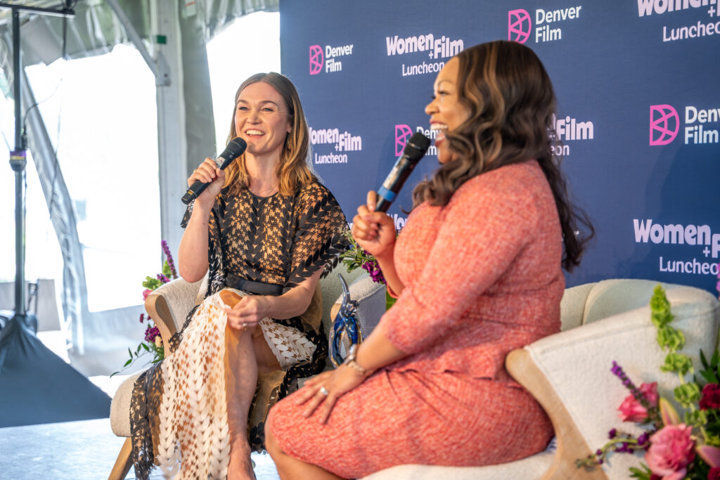 Two women with microphones at Women+Film Luncheon event, smiling and seated on stage