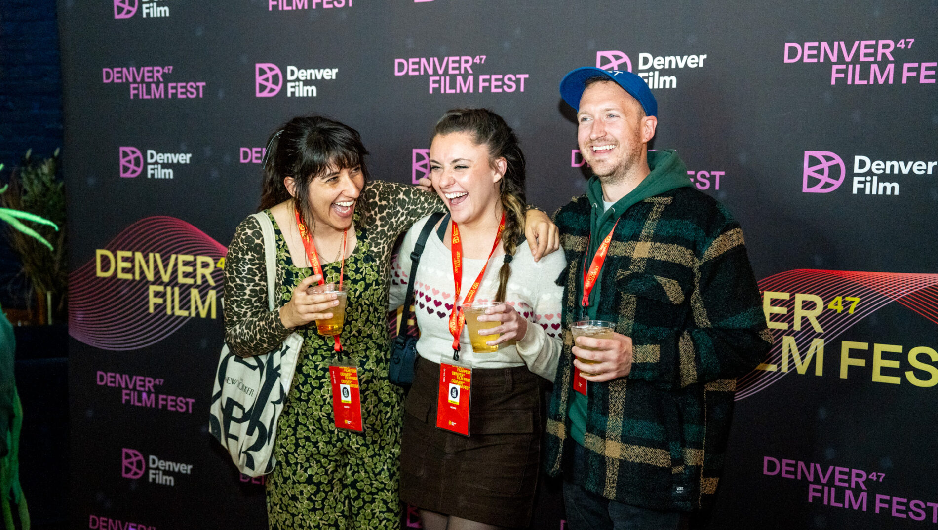 Three people laughing and holding drinks in front of Denver Film Fest backdrop