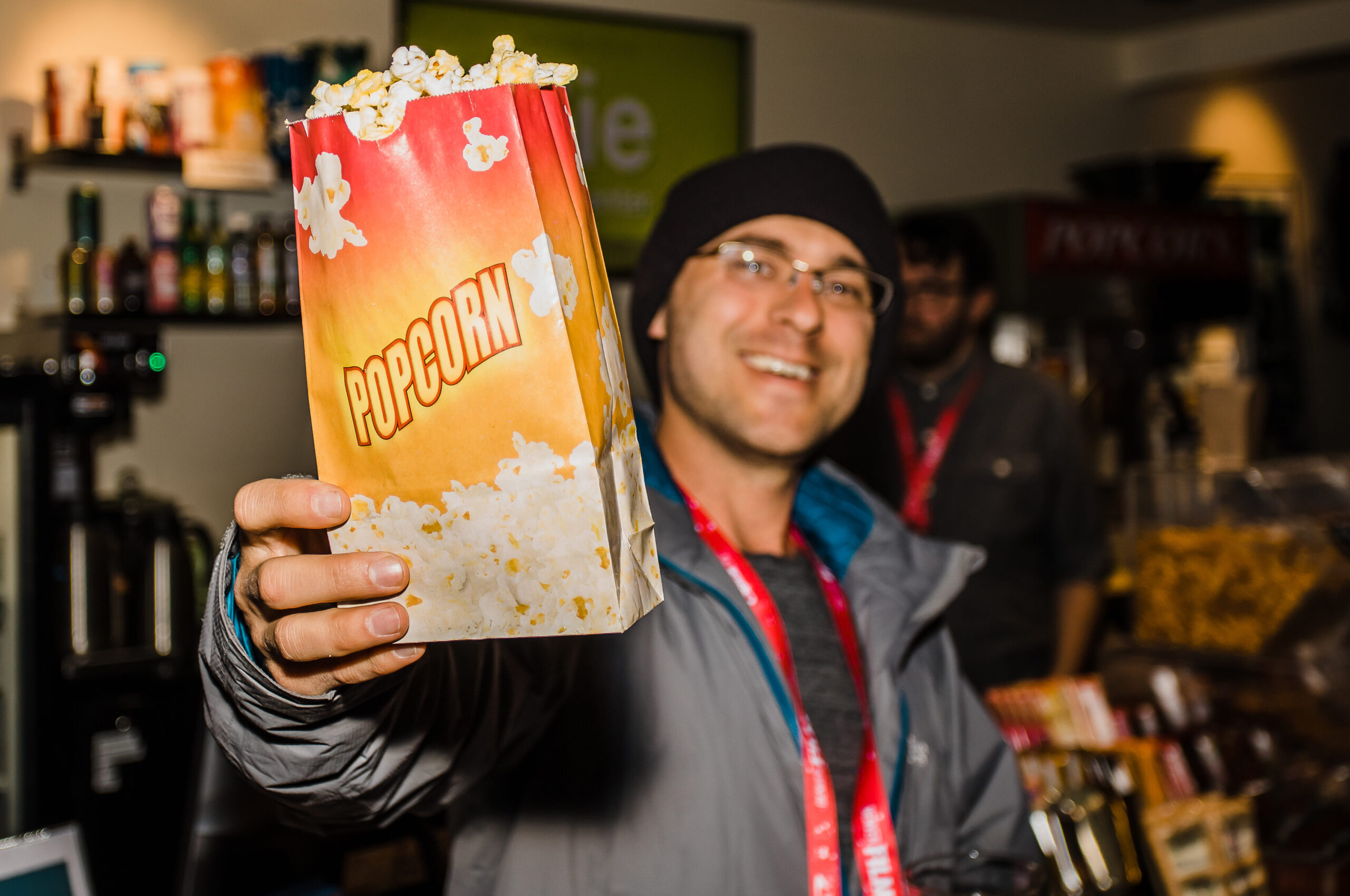 Person smiling, offering popcorn in a concession area, wearing a beanie and glasses