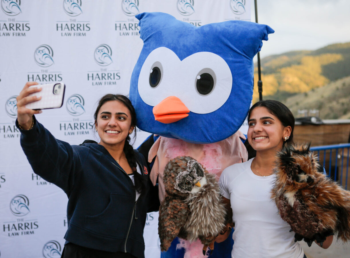 Two smiling women taking a selfie with a mascot in a blue bird costume in front of "The Harris Law Firm" backdrop