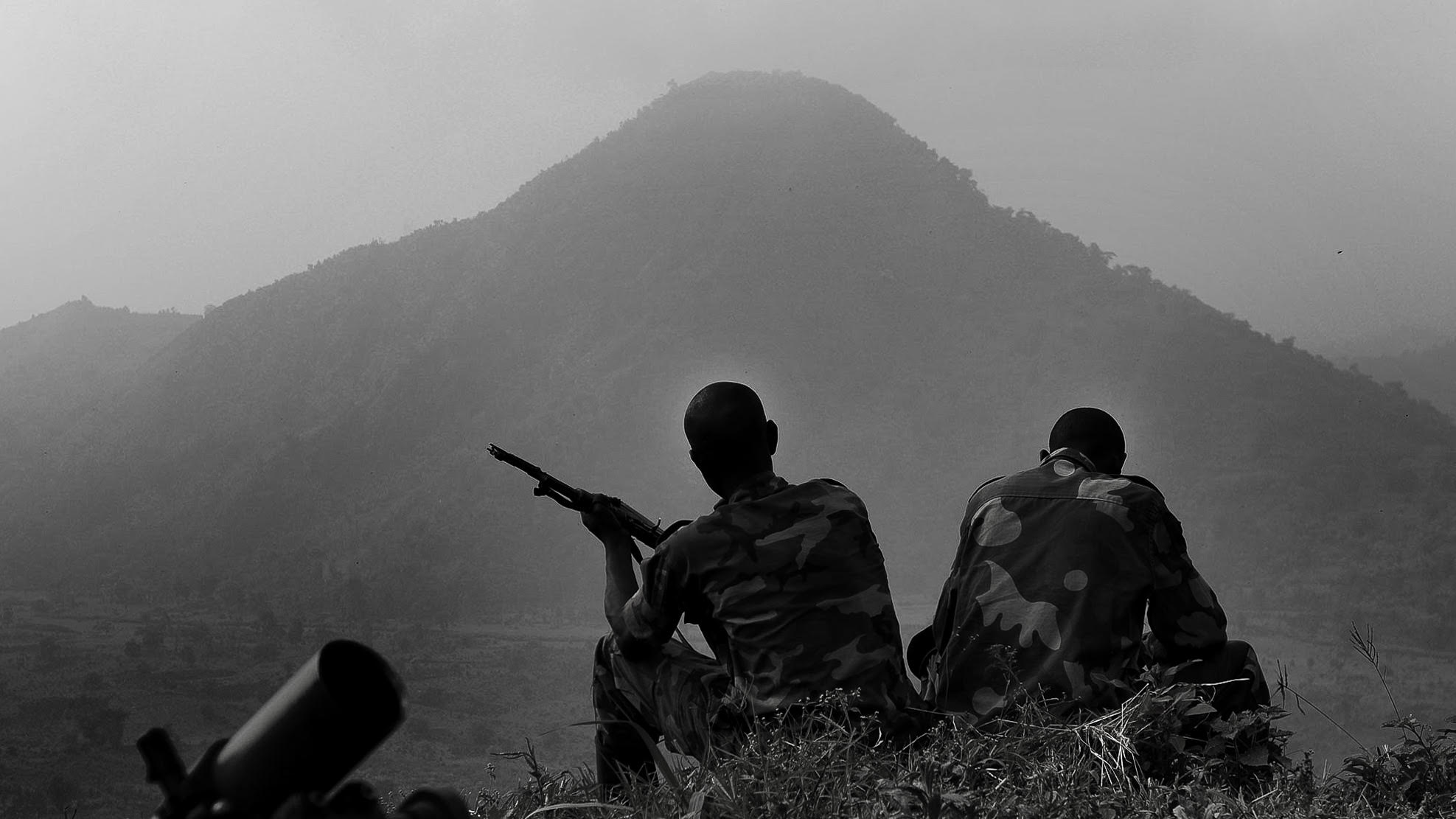 Two soldiers in camouflage sit on a hill, one holding a rifle, overlooking a misty mountain landscape