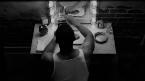 A man in a tank top sits at a vanity table with bright lights, facing a mirror displaying his reflection