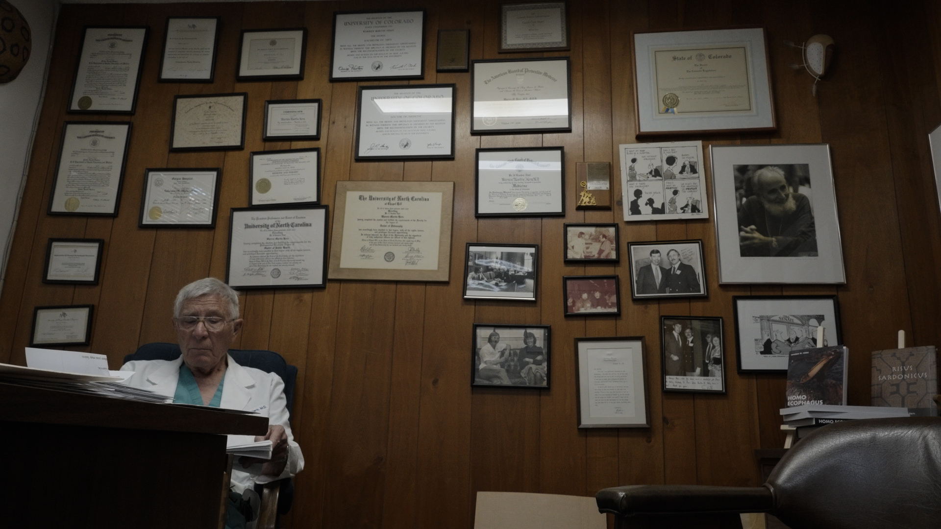 Man in white coat sits in office with numerous diplomas and photos on wood-paneled wall