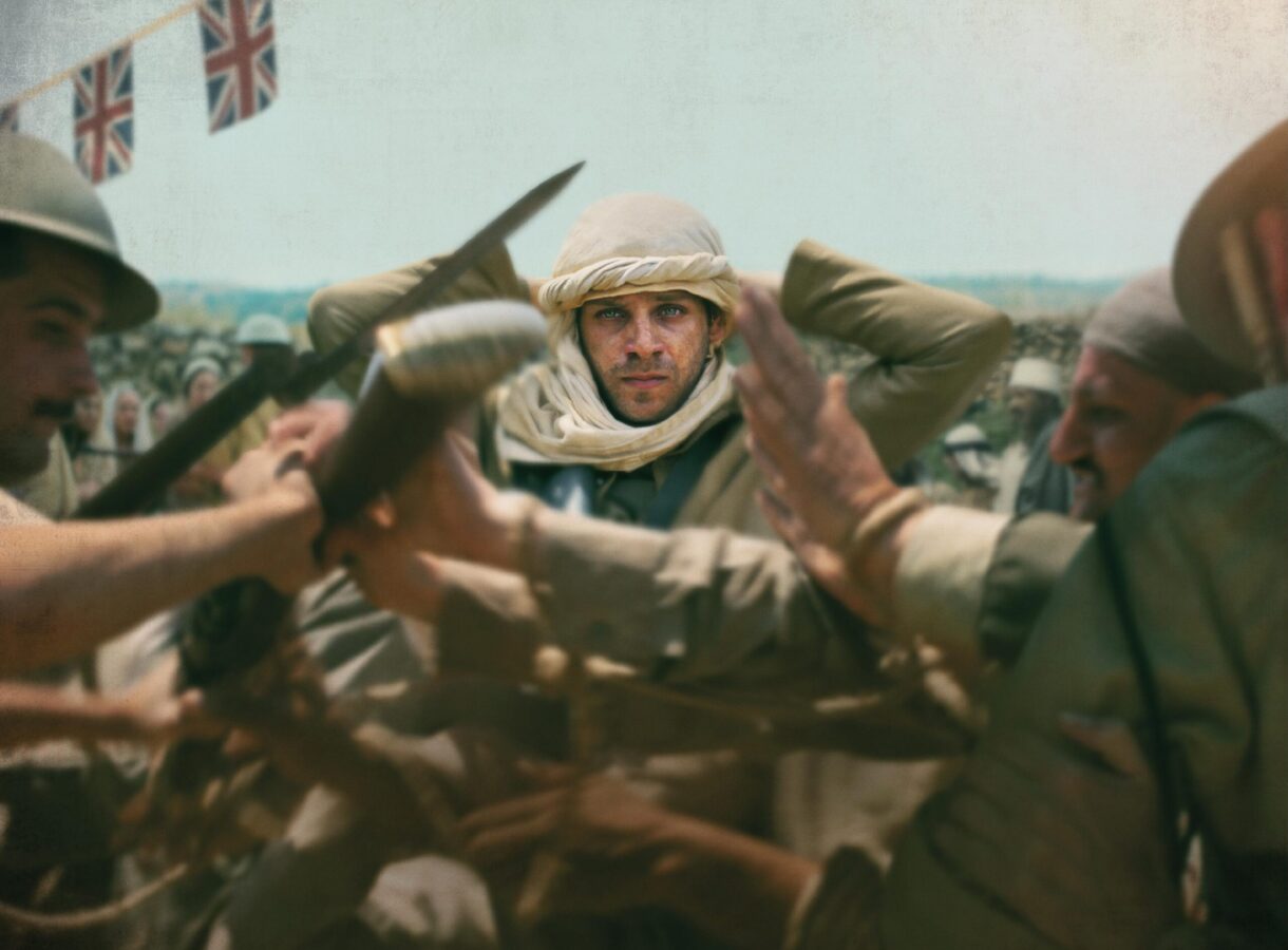 A man in traditional attire stands with hands raised amidst a tumultuous crowd, British flags overhead, intense expression