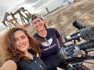 Two women smiling near oil pumps, one holding a camera. The woman in the "Meowhaus" shirt wears a backward cap