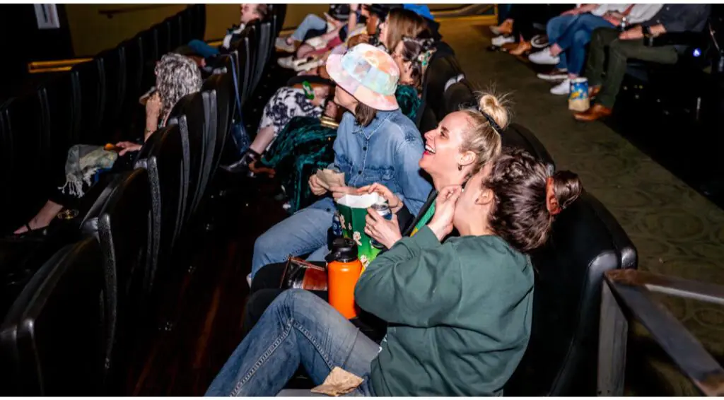 [gpt-4o]: Three people sitting in theater seats, smiling and holding drinks, surrounded by other attendees