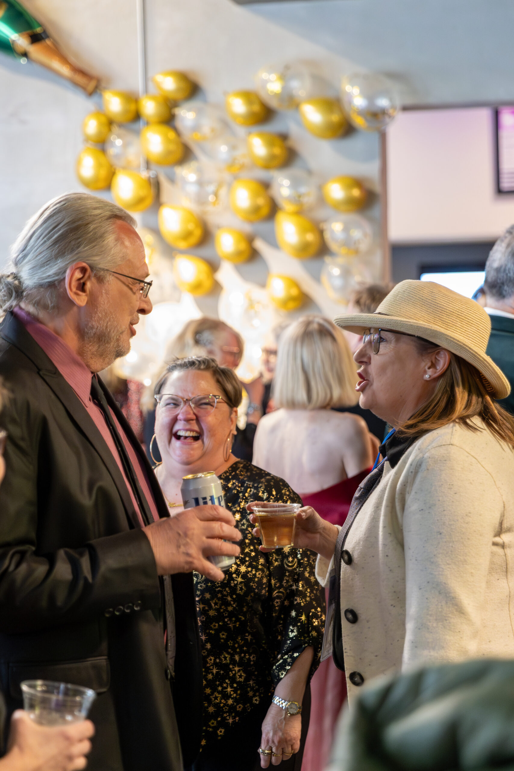 [gpt-4o]: People socializing at a gathering, holding drinks, with gold and clear balloons on the wall in the background